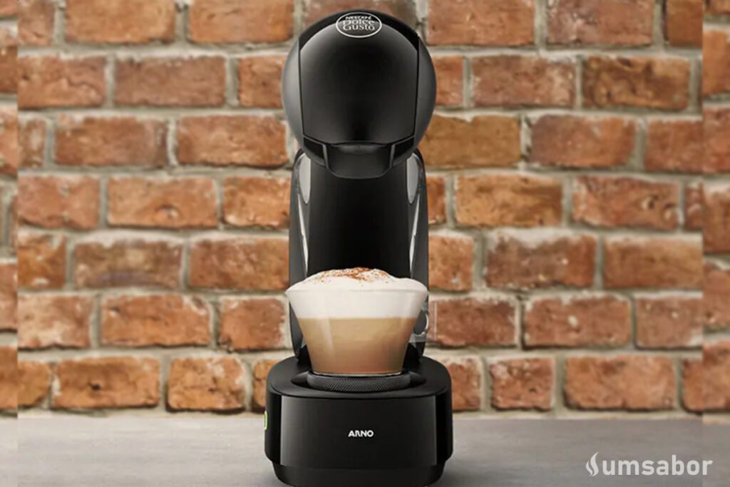 A imagem contém a cafeteira de cápsula dolce gusto infinissima ao centro com um copo com bebida a base de leite sendo preparada nela. o restante do fundo é uma parede de tijolos expostos.