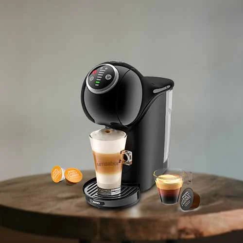 A imagem é composta por uma mesa de madeira e em cima dessa mesa há, da esquerda para a direita, duas cápsulas da dolce gusto, uma cafeteira dolce gusto genio s plus de cor preta com um copo com capuccino dentro,  um copo com café e uma cápsula de café.