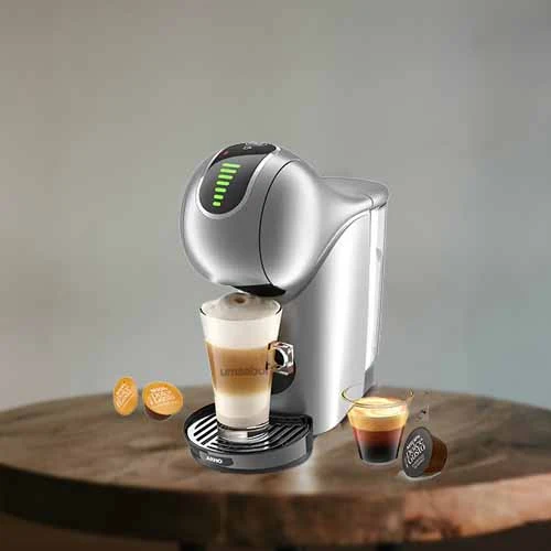 A imagem é composta por uma mesa de madeira e em cima dessa mesa há, da esquerda para a direita, duas cápsulas da dolce gusto, uma cafeteira dolce gusto genio s touch de cor prata com um copo com capuccino dentro,  um copo com café e uma cápsula de café.