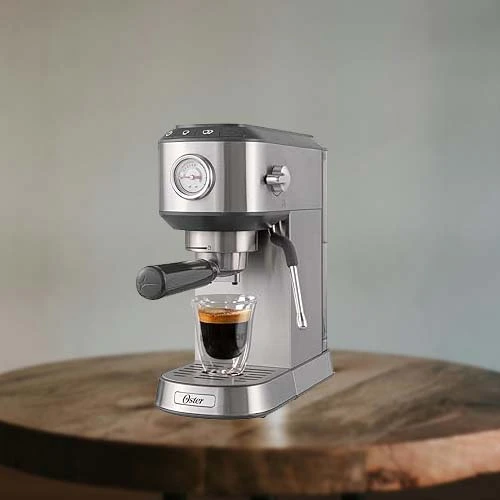 A imagem é composta por uma mesa de madeira e em cima dessa mesa há uma cafeteira de modelo Compact Perfect Brew da marca Oster de cor prata com um copo com café dentro