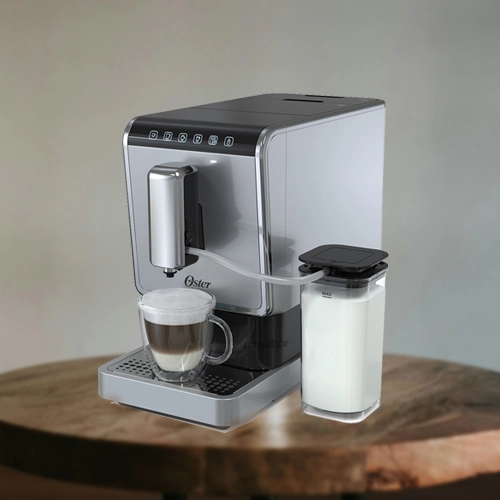 A imagem é composta por uma mesa de madeira e em cima dessa mesa há uma cafeteira de modelo superautomatica da marca Oster de cor prata com um copo com café dentro e o reservatório de leite ao lado abastecido