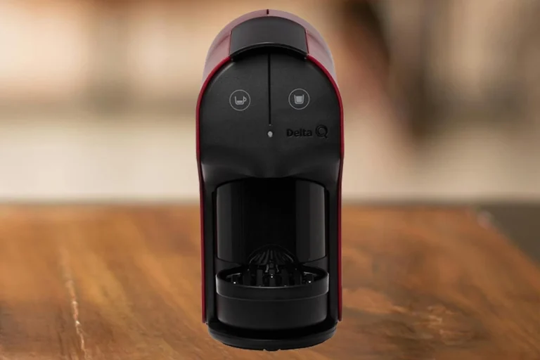 Delta Q Quick é boa? Vale a pena comprar em 2026? 1 A imagem contém a cafeteira de cápsula Delta Q Quick na cor preta com vermelho. Essa cafeteira está ao centro da imagem e em cima de uma mesa de madeira com o fundo desfocado.