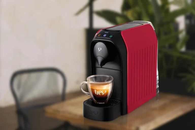Três Corações Passione: desempenho, funcionalidades e vale a pena comprar? 1 Trata-se de uma imagem que contém uma cafteira de cápsula de cor vermelha com preto e o fundo da imagem há uma mesa, plantas e cadeira. Essa cafeteira é a Três Corações Passione