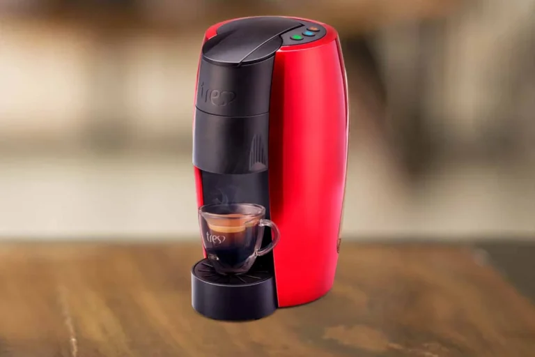 Três Corações Lov: análise técnica e review com opinião especializada 8 A imagem é composta pela cafeteira de cápsula Três Corações Lov de cor preta com vermelho ao centro da imagem. Essa cafeteira está sob uma mesa de madeira e o restante do fundo é desfocado. Há nessa cafeteira uma xícara com café expresso.