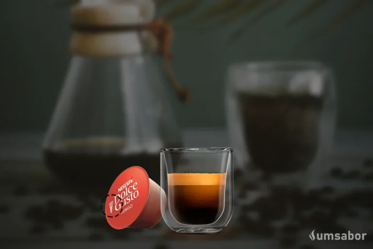 Uma cápsula de café Dolce Gusto Lungo vermelha ao lado de um copo de vidro transparente com café espresso e crema, com grãos de café ao fundo.